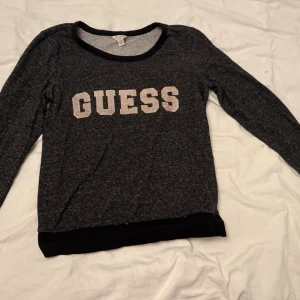 Mörkgrå långärmad tröja från Guess - Säljer en mörkgrå långärmad tröja från Guess i storlek XS. Tröjan har ett stort vitt Guess-tryck på bröstet och svarta ribbade detaljer vid hals och nederkant. Perfekt för dig som gillar enkel och snygg streetstil.