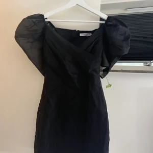 Svart kort klänning från Adoore - Supersnygg svart klänning från Adoore i kort modell med puffiga axlar och omlott-v-ringning. Klänningen har en elegant och modern siluett som verkligen sticker ut. Perfekt för dig som vill ha något stilrent med en cool twist. Klänningen säljs inte längre på deras sida och har endast används ett fåtal gånger, perfekt skick, orginalpris på klänningen ligger runt 600kr 