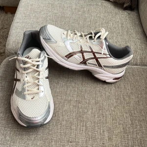 Asics Gel-1130 Cream Reddish/Brown - Snygga vita Asics sneakers med silvriga och vinröda detaljer. Ovandelen är i mesh och syntet för bra andningsförmåga och komfort. Skorna har snörning, grå insida och en platt sula som passar perfekt till en sportig look.
