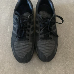 Svarta Adidas LA Trainer sneakers - Snygga svarta Adidas LA Trainer sneakers med klassiska tre ränder på sidan. Ovandelen är i mesh och syntet för en sportig look och bra andningsförmåga. Skorna har snörning och platt sula, perfekt för en aktiv vardag.