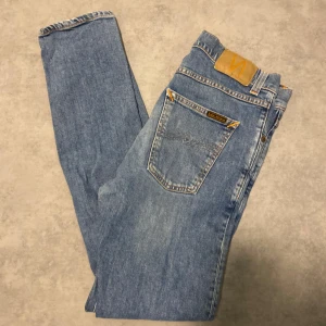 Nudie lean Dean jeans - Säljer ett par klassiska ljusblå jeans från Nudie Jeans med raka ben och snygga slitningar. Jeansen har dragkedja, orange kontrastsömmar och Nudie-logga på bakfickan. Perfekta för en avslappnad och stilren look. Jeansen har två defekter ena är att dragkedjan behöver lagas samt så är de sydda i skrevet men inget som märks vid användning😁