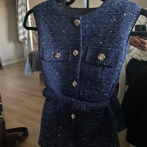 Mörkblå glittrig väst med bälte - Snygg mörkblå väst i glittrigt tyg med guldiga knappar framtill och två fickor på bröstet. Västen har ett matchande bälte i midjan och är ärmlös med rund halsringning. Perfekt för att lyfta vilken outfit som helst.