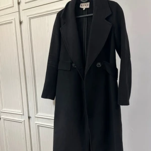 Svart lång kappa från NA-KD - Stilren svart kappa från NA-KD med klassisk krage och knappar framtill. Lång modell med fickor på sidorna och snyggt skräddad passform. Perfekt för dig som gillar en clean och tidlös look.