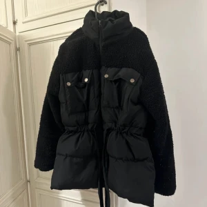 Svart teddy- och pufferjacka med snörning - Snygg svart jacka som kombinerar teddy- och pufferstil. Hög krage, dragkedja framtill och praktiska fickor med knappar. Jackan har snörning i midjan för en cool siluett och är perfekt för kyliga dagar.