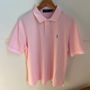Rosa pikétröja från Polo Ralph Lauren - Snygg rosa pikétröja från Polo Ralph Lauren med klassisk krage och korta ärmar. Tröjan har blå broderad logga på bröstet och två knappar vid halsen. Tillverkad i mjuk bomull som är skön mot huden. Perfekt för dig som gillar stilren och fräsch look.