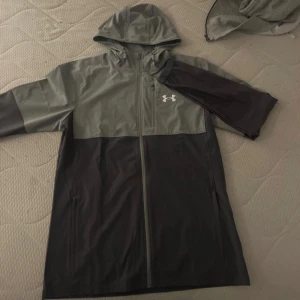 Under Armour vindjacka grå/svart - Säljer en snygg tvåfärgad vindjacka från Under Armour i grått och svart. Helt oanvänd skick 10/10Jackan har huva med snörning, dragkedja hela vägen och en bröstficka med dragkedja. Perfekt för blåsiga dagar och sportiga outfits.