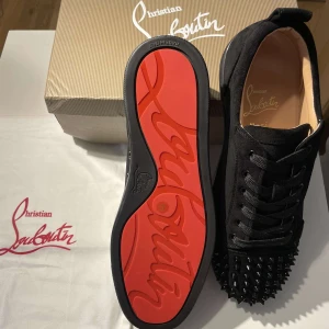 Christian Louboutin svarta sneakers med nitar - Svarta sneakers från Christian Louboutin i mocka med ikonisk röd sula och svarta nitar på tån. Skorna har snörning och rund tå, vilket ger en edgy och lyxig vibe. Perfekta för dig som vill sticka ut med exklusiva detaljer och streetstyle-känsla.