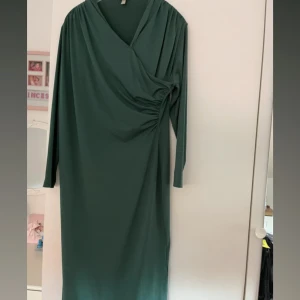 Grön omlottklänning från H&M - Mörkgrön långklänning från H&M med omlott-design och rynkade detaljer vid sidan. Klänningen har lång ärm och v-ringad hals. Tillverkad i mjukt polyester-tyg som faller snyggt och känns bekvämt. Hann aldrig använda den så helt oanvänd!