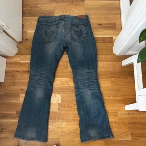 Bootcut jeans med svan/hjärt detalj - Säljer ett par blå bootcut jeans med snygg tvätt och hjärtformade detaljer på bakfickorna. Jeansen har klassisk femficksdesign, knapp- och dragkedjegylf samt lätt slitna partier för en cool look. Perfekta för dig som gillar retrovibbar och unik stil.