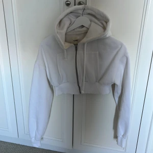 Vit croppad hoodie från Pull&Bear - Säljer en vit croppad hoodie från Pull&Bear i storlek S. Tröjan har dragkedja framtill, huva med snörning och långa ärmar. Perfekt för en chill och avslappnad stil. Tillverkad i mjukt material som känns skönt mot huden.