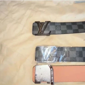 Louis Vuitton svart skärp med LV-spänne - Snyggt svart Louis Vuitton-bälte i skinn med klassiskt monogrammönster och stort silverfärgat LV-spänne. Bältet har en bred design och diskreta gråa detaljer i mönstret. Perfekt för att lyfta vilken outfit som helst med en lyxig touch.