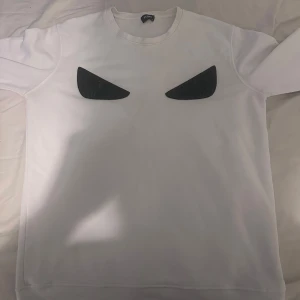 Vit sweatshirt från Fendi med ögon - Säljer en vit sweatshirt från Fendi i storlek XL men passar L med ikoniska svarta ögon på bröstet. Tröjan har rund halsringning och långa ärmar, perfekt för dig som vill sticka ut med en cool och stilren design.