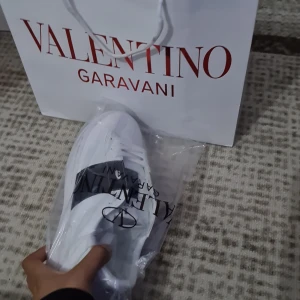 Valentino Rockstud vita sneakers - Snygga vita Valentino Rockstud sneakers med svart band och ikoniska nitar över vristen. Skorna har rund tå, platt sula och klassisk snörning. Tillverkade i skinn för en lyxig känsla och stilren look. Perfekta för dig som vill sticka ut med exklusiva detaljer.
