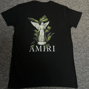 Svart Amiri t-shirt med ängeltryck - Svart t-shirt från Amiri med stort tryck av en vit ängel och gröna blad på ryggen samt mindre tryck på bröstet. Rund hals och korta ärmar. Materialet är mjuk bomull och passformen är normal. Snygg kontrast mellan svart tyg och detaljerna i trycket.
