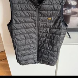 Svart väst Barbour M - Säljer en svart quiltad dunväst från Barbour International. Västen har en stilren look medp, dragkedja framtill och en liten gul Barbour-logga på bröstet. 