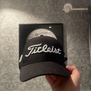 Svart Titleist keps Pro V1 - Svart keps från Titleist med broderad logga framtill och texten PRO V1 på sidan. Kepsen har även FJ-brodyr på sidan och ett patch med röd kant bak. Tillverkad i ett strukturerat, lätt material som andas. Perfekt för golfbanan eller sportig stil. I storlek L/XL