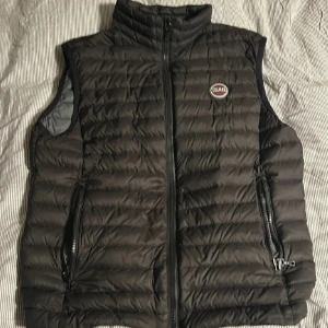 Svart dunväst från Colmar - Säljer en svart dunväst från Colmar med quiltad design och hög krage. Västen har två fickor med dragkedja och Colmar-logga på bröstet. Perfekt att slänga över en hoodie eller tröja när det är kyligt ute. Stängs med dragkedja framtill.