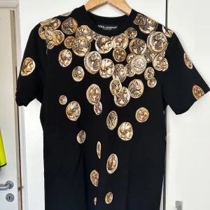 Svart t-shirt med guldmynt från Dolce & Gabbana - Svart t-shirt från Dolce & Gabbana med unikt tryck av antika guldmynt över hela plagget, både fram och bak. Klassisk rund halsringning och korta ärmar. Snygg statement-piece med lyxig vibe, perfekt för dig som vill sticka ut.