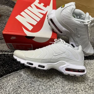 Nike Air Max Plus vita/grå sneakers - Nike Air Max Plus i fräscht vitt och grått med ikoniska vågiga detaljer och synlig Air Max-dämpning. Ovandel i mesh och syntet, robust sula med svarta och röda detaljer undertill. Perfekta för dig som vill sticka ut med en klassisk streetwear-look.