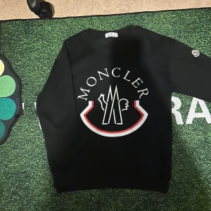 Svart Moncler sweatshirt med logga - Svart sweatshirt från Moncler med stor vit och röd logga framtill och broderad patch på ärmen. Klassisk rund halsringning och lång ärm. Perfekt för dig som gillar streetwear och vill sticka ut med en ikonisk look.