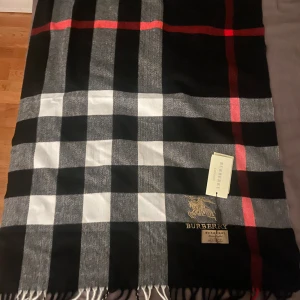 Rutig Burberry halsduk i kashmir - Snygg halsduk från Burberry i 100% kashmir med klassiskt rutigt mönster i svart, grått, vitt och röda detaljer. Broderad Burberry-logga i guld och fransar i ändarna. Aldrig använd vet inte vart låda och kvitto är därför billigt pris.