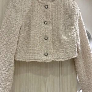 Beige croppad kavaj med pärlknappar - Snygg croppad kavaj i beige med strukturerat tyg och pärlknappar framtill. Jackan har långa ärmar, råa kanter och en klassisk, elegant look. Perfekt för dig som vill ha en chic och trendig stil med en twist.