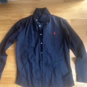 Mörkblå skjorta från Ralph Lauren - Klassisk mörkblå skjorta från Ralph Lauren med lång ärm och knappar framtill. Skjortan har den ikoniska röda logotypen broderad på bröstet och är tillverkad i bomull. Perfekt för dig som gillar stilrena och tidlösa plagg.