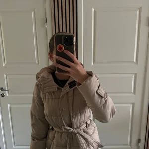Beige pufferjacka med bälte - Säljer en beige pufferjacka med huva och knytbälte i midjan. Jackan har långa ärmar och quiltad design som ger en riktigt mysig vibe. Perfekt för kyliga dagar när du vill vara både varm och snygg.