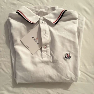 Vit Moncler pikétröja med röd/blå kant - Vit pikétröja från Moncler med klassisk krage och korta ärmar. Tröjan har snygga rödblå ränder på krage och ärmslut samt Moncler-logga broderad på bröstet. Tillverkad i mjuk bomull och har knappar framtill. Perfekt för en clean och sportig look.                                                                                                                                                Nypris: 3600kr GORILLAPRIZE:1820kr            Skick:9/10 testad                           KVITTO MEDKOMMER     
