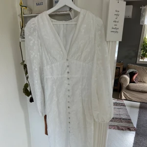 Vit långklänning med broderade blommor - Superfin vit långklänning med v-ringning och puffiga långärmar. Klänningen har broderade blommor över hela tyget och knappar framtill. Materialet känns lätt och luftigt, perfekt för sommarens alla tillfällen. Dragkedja i ryggen för enkel påklädning.