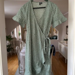 Grön prickig omlottklänning Vero Moda - Snygg grön omlottklänning från Vero Moda med vita prickar och knyt i midjan. Klänningen har korta ärmar, v-ringad hals och volangdetalj nedtill. Perfekt för sommarens alla tillfällen och superenkel att styla med sneakers eller sandaler.