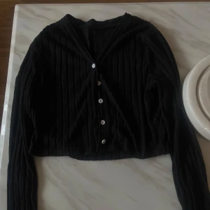 Svart ribbad kofta med v-ringning - Svart ribbad kofta med v-ringning och vita knappar framtill. Croppad modell med långa ärmar, perfekt att slänga över en topp eller klänning. Enkel och stilren design som funkar till många outfits.