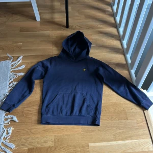Mörkblå hoodie från Lyle & Scott - Säljer en mörkblå hoodie från Lyle & Scott med klassisk gul logga på bröstet. Tröjan har huva, känguruficka och långa ärmar. Perfekt för dig som gillar en enkel och stilren look. Materialet är mjuk bomull som känns skönt mot huden.