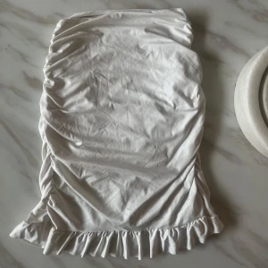 Vit kortkjol med volangkant - Supersöt vit kortkjol med volangkant längst ner och snygga rynkade detaljer längs sidorna. Kjolen har en feminin och fräsch look, perfekt för dig som gillar ljusa och stilrena plagg. Materialet känns mjukt och lätt mot huden.