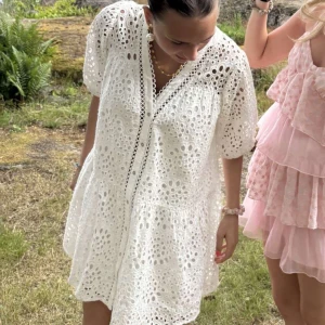 Vit kort klänning med broderade hål - Superfin vit klänning med broderade hål och puffiga kortärmar. Klänningen har en luftig passform, v-ringad hals och volang nedtill. Perfekt för sommarens alla tillfällen, och materialet känns som bomull. Söt och trendig look!