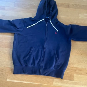 Mörkblå hoodie från Polo Ralph Lauren strl M  - Snygg mörkblå hoodie från Polo Ralph Lauren med klassisk röd logobroderi på bröstet. Tröjan har huva med vita snören, dragkedja framtill och två fickor. I nyskick då den inte använts väldigt sparsamt. 