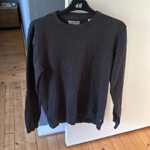 Mörkgrå stickad tröja Jack & Jones - Snygg mörkgrå stickad tröja från Jack & Jones i storlek M. Tröjan har rund hals, långa ärmar och ribbade muddar vid ärmslut och nederkant. Perfekt för dig som gillar enkel och stilren design med en diskret detalj vid nederkanten. Inte använd så mycket, så tröjan är i väldigt fint skick!