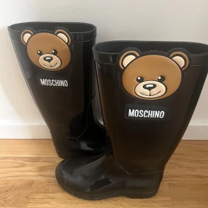 Svarta Moschino gummistövlar med björn - Coola svarta gummistövlar från Moschino med stora björndetaljer framtill och tydlig logga. Stövlarna är tillverkade i blankt syntetmaterial och har en klassisk hög modell som sticker ut med sin lekfulla design.