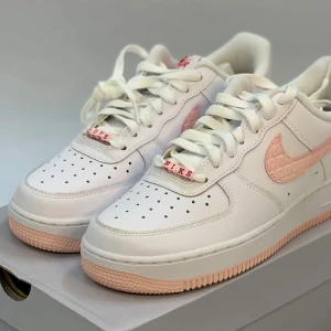 Nike Air Force 1 Low Valentine’s Day 2022 - Nike Air Force 1 sneakers i vitt skinn med ljusrosa detaljer på swoosh och yttersula. Skorna har snörning med vita skosnören och små röda bokstäver på lace tags. Perforerad tå för extra ventilation och klassisk rund tå. Perfekt för dig som gillar clean och fräsch stil.