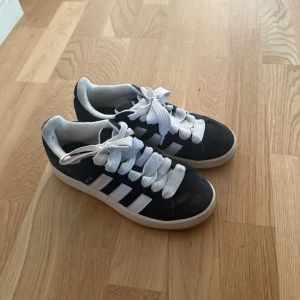 Adidas Campus sneakers svart/vit - Snygga Adidas Campus sneakers i svart mocka med klassiska vita ränder och vit sula. Skorna har breda vita snören och gummisula i beige. Perfekta för dig som gillar retrostil och streetwear.