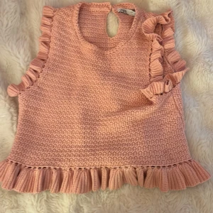 Stickad rosa volangtopp från Zara - Supersöt stickad topp från Zara i ljusrosa med volangdetaljer längs ärmhål och nederkant. Ärmlös modell med rund hals och knappslå i nacken. Perfekt för dig som gillar en gullig och trendig look.