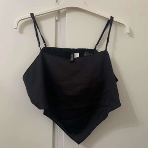 Svart bandana-linnetopp från Gina Tricot - Snygg svart linnetopp från Gina Tricot med bandanaformad nederkant och tunna justerbara axelband. Perfekt för dig som vill ha en enkel men trendig topp till sommaren. Toppens material känns mjukt och lätt mot huden.