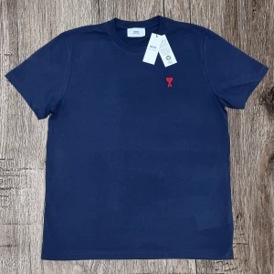 Marinblå t-shirt från Ami med hjärtdetalj - Snygg marinblå t-shirt från Ami med klassisk passform och korta ärmar. På bröstet finns ett broderat rött hjärta med Ami-loggan. Tillverkad i mjuk bomull som känns skön mot huden. Perfekt för dig som gillar stilrena plagg med en twist. Storlek M