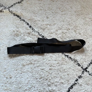 Svart syntetbälte med plastspänne - Säljer ett svart bälte i syntetmaterial med ett rejält plastspänne. Bältet är slätt och har en enkel, funktionell design som passar till streetwear eller outdoor. Perfekt för dig som gillar praktiska accessoarer med minimalistisk stil.