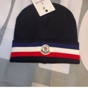 Svart Moncler mössa med ränder - Svart mössa från Moncler med klassiska blå, vita och röda ränder på uppviket. Framtill sitter Monclers logotyp. Perfekt accessoar för att lyfta din vinterstil.
