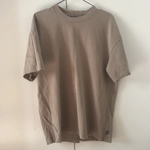 Beige t-shirt med ribbad krage - Snygg beige t-shirt med ribbad rund halsringning. Modellen är enkel och har en loose passform med korta ärmar. Perfekt för dig som gillar stilrena och neutrala plagg.