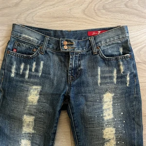 Blå distressed jeans från Seven7 - Säljer ett par blå jeans från Seven7 med coola slitningar och broderade bakfickor. Jeansen är utsvängda, dubbla knappar i midjan och tydliga  detaljer framtill. Använt få gånger, pris kan även diskuteras.
