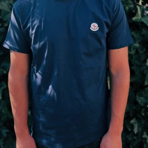 Mörkblå Moncler t-shirt i bomull - Snygg mörkblå t-shirt från Moncler med klassisk rund hals och diskret logotyp på bröstet. Tillverkad i mjuk bomull som känns skön mot huden. Passformen är normal och den har korta ärmar. Perfekt för dig som gillar stilrena plagg med exklusiv touch.                                                                                                                                                                              Nypris: 2000kr GORILLAPRIZE:1120kr     Skick:10/10 oöppnad   KVITO MEDKOMMER
