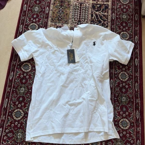 Vit Polo Ralph Lauren t-shirt - Vit t-shirt från Polo Ralph Lauren med klassisk pikékrage och korta ärmar. T-shirten har den ikoniska broderade loggan i marinblått på bröstet och är tillverkad i mjuk bomull. Perfekt för en clean och stilren look.