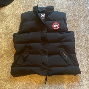Svart dunväst från Canada Goose - Svart dunväst från Canada Goose med klassisk logga på bröstet. Västen har hög krage, tryckknappar framtill och två fickor med lock. Perfekt för lager-på-lager och riktigt varm tack vare dunfyllningen. Snygg och stilren design som funkar till det mesta. Strl xl men passar mindre strl med